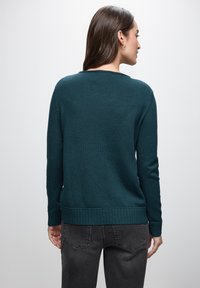 Maglione a maglia turchese con scollo rotondo, polsini e orlo a coste, abbinato a jeans slim-fit grigio scuro. Texture liscia, dettagli minimali.