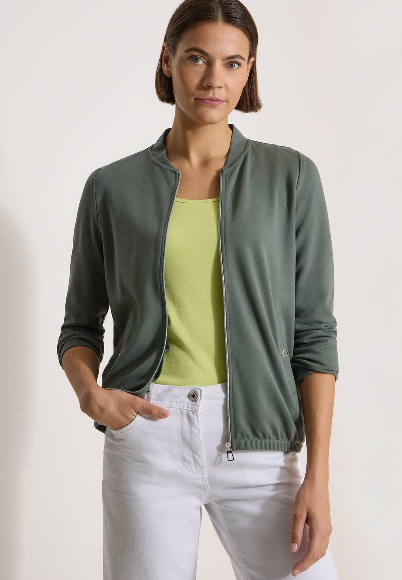 Femme portant une veste verte zippée par-dessus un haut vert clair, avec un pantalon blanc, debout devant un fond uni.