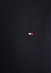 Černá pletená tkanina s malým, trojbarevným logem v červené, bílé a modré barvě našitým na levé straně. Žádné další pozoruhodné detaily.