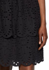 Robe noire avec un design en couches brodé présentant des motifs découpés et un ourlet festonné. Le matériau semble texturé avec une silhouette structurée.
