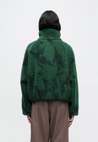 Πράσινο φλις μπουφάν με ψηλό γιακά, υφή στην επιφάνεια και άνετη εφαρμογή. Το πουλόβερ φαίνεται μαλακό, με κοντό σχεδιασμό και χαμηλωμένους ώμους.