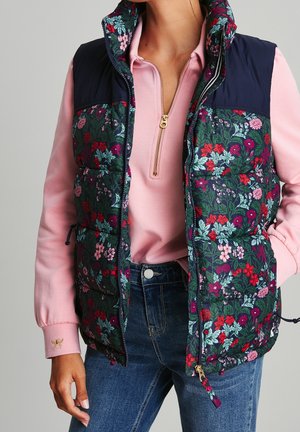 Kobieta w pikowanej kamizelce w kwiatowy wzór na różowym swetrze z krótkim suwakiem i niebieskich dżinsach, z ręką spoczywającą blisko kieszeni.