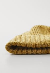 Cască din lână tricotată, galben muștar, pliată, cu textură ribbed, pe o suprafață albă.