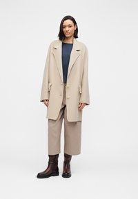 Beige wool coat with large lapels and two buttons, worn over a navy top and beige wide-leg pants, paired with brown ankle boots.