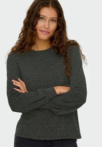 Zwarte trui met een gestructureerd, ruitvormig breipatroon. Ronde hals, lange mouwen en een losse pasvorm. Armen gekruist tegen een effen achtergrond.