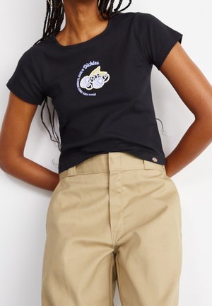 Žena oblečená v černém krátkém tričku Dickies s grafickým potiskem a béžových kalhotách s vysokým pasem, stojící před bílým pozadím.