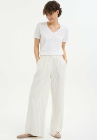 Witte v-hals t-shirt gecombineerd met beige gestreepte wijde broek met knopen aan de voorkant en zijzakken. Licht schoeisel maakt de outfit compleet.