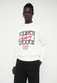 Biała sweterkowa bluzka z wielokolorowym logo "TOMMY JEANS" z przodu. Ściągacze przy mankietach i na dolnej krawędzi, o gładkiej strukturze. Noszona z czarnymi spodniami.