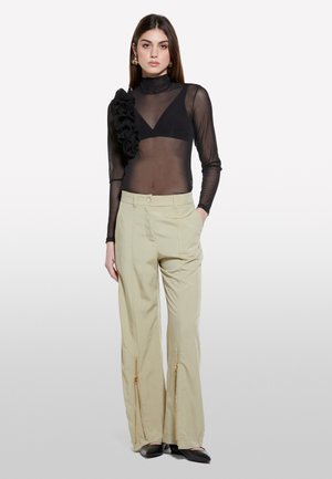 Bodysuit noir transparent avec épaule gauche à volants ; associé à un pantalon large beige avec des accents de fermeture éclair à l'avant. Chaussures plates noires.