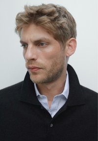 Schwarzer Strickpullover mit umgeschlagenem Kragen, über einem hellblauen und weißen gestreiften Hemd getragen. Strukturierter Stoff und subtile Nähdetails.