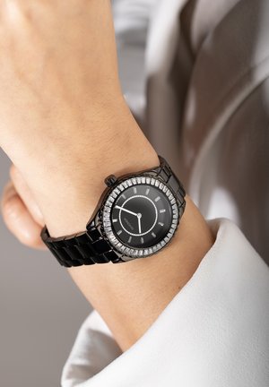 Montre noire avec un bracelet brillant et un cadran rond, featuring un cadran noir, des index argentés et une lunette ornée de cristaux.