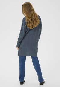 Femme aux longs cheveux châtain clair portant un cardigan en tricot gris, un jean bleu et des chaussures noires, se tenant devant un fond blanc uni.