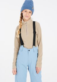 Beige fleece zip-up tröja med svarta hängslen, tillsammans med ljusblå snöbyxor med sidofickor och en avslappnad passform. En blå stickad mössa fulländar looken.