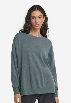 Jasnoniebiesko-zielony, oversized sweter z długimi rękawami, prążkowanym dekoltem i dołem. Miękka faktura i luźny krój, w połączeniu z ciemnymi dżinsami.