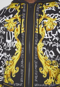 Sweat à capuche noir zippé avec des motifs baroques dorés et un texte graffiti blanc incluant "Versace Jeans Couture" sur les panneaux avant.