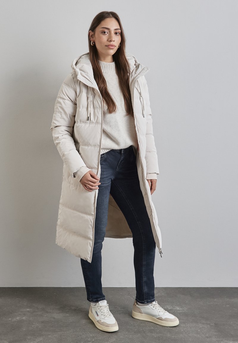 Lång beige pufferjacka med huva, sidofickor och ett quiltat mönster, kombinerad med en ljus tröja och mörka jeans. Vita sneakers fulländar looken.