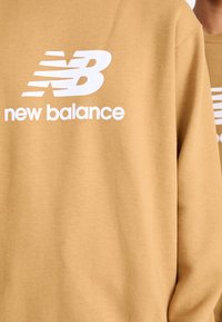 Sudadera color arena con un audaz logo en blanco de "New Balance" y líneas diagonales de acento. El material parece suave y tiene una textura lisa.