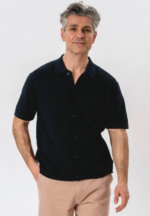 Stredoveký muž so sivými vlasmi, oblečený v čiernu krátkym rukávom zapínanú košeľu a béžové nohavice, stojaci s jednou rukou v vrecku.