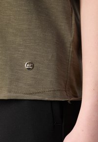 Olijfgroene, getextureerde T-shirt met een kleine zilveren logo-badge nabij de zoom en contrasterende zwarte broek. Naaddetails zichtbaar aan de rand.