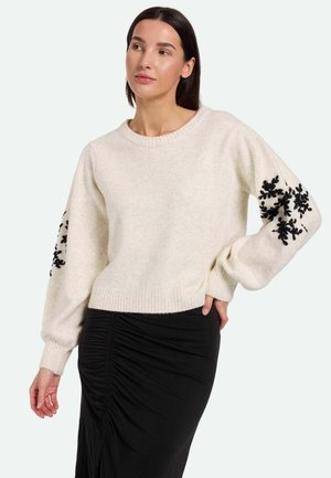 Pull en laine crème avec broderie florale noire sur les manches. Encolure ronde et ourlet côtelé. Associé à une jupe noire ajustée et froncée.