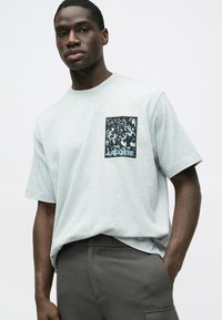 Jasnoszara koszulka z miękkiego materiału z czarną kieszenią w kształcie kwadratu ozdobioną niebiesko-białym wzorem. Model nosi ciemnoszare spodnie.