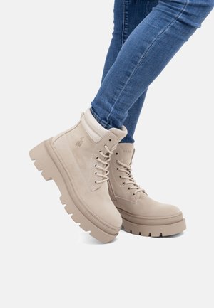 Stivaletti alla caviglia in pizzo beige chiaro con suole spesse indossati con jeans skinny blu su uno sfondo bianco.