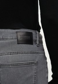 Primer plano del bolsillo trasero de unos vaqueros de mezclilla grises con una etiqueta de cuero negro grabada con la palabra "DRYKORN", combinados con una camiseta negra.