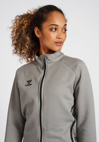 Lys grå zip-up jakke med høj krave, tekstureret stof, sorte lynlåslommer og et lille logo på venstre bryst.