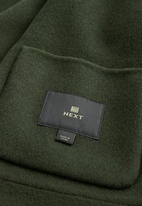 Mørkegrønt fleece-stof med en lomme og en sort label med guld "NEXT" logo. Blød tekstur, glat finish.