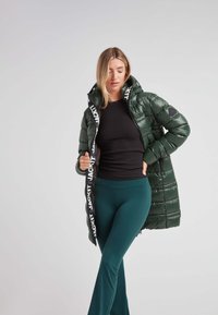 Chaqueta acolchada verde con un acabado brillante, que presenta un cuello alto y una marca blanca en la cremallera. El modelo lleva una camiseta negra y pantalones en tono verde azulado.