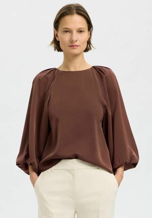 Blusa marrone fluida con scollatura rotonda, maniche a sbuffo e texture liscia; abbinata a pantaloni su misura di colore chiaro.