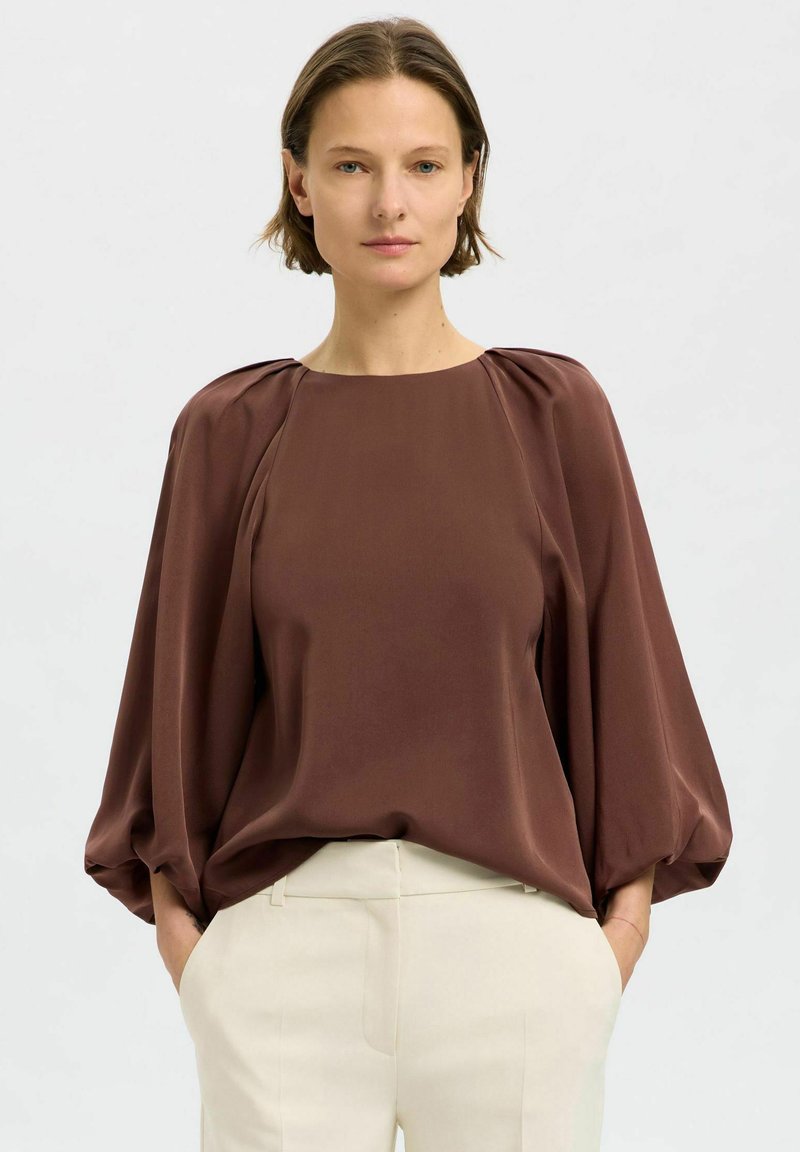 Blusa marrone fluida con scollatura rotonda, maniche a sbuffo e texture liscia; abbinata a pantaloni su misura di colore chiaro.