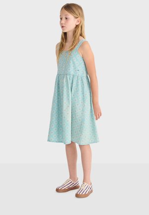 Jeune fille portant une robe sans manches bleu clair à motifs et des baskets rayées, debout et de profil sur un fond uni.