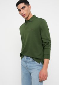 Zelená dlhá polo košeľa s golierom, s malým logom na hrudi. Nosená s svetlomodrými džínsami, ktoré majú voľný strih.