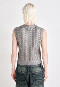 Veste sans manches grise en tricot avec motif torsadé, col et ourlet côtelés. Portée avec un jean taille haute, présentant des détails de texture et de poche.