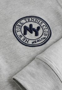 Hall müts, millel on ümmargune tumesinine tikandiga logo "New York Tennis Club". Omab pehmet tekstuuri ja mansetiga varrukaid.