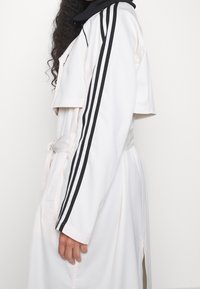 Vit trenchcoat med svarta ränder längs ärmarna, med skärp i midjan och sidospjäll. Slät tyg och strukturerad design.