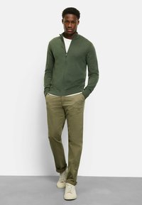 Grüne Reißverschluss-Strickjacke aus weichem Material, kombiniert mit helloliven Chinos und cremefarbenen Sneakers. Das Modell steht vor einem neutralen Hintergrund.