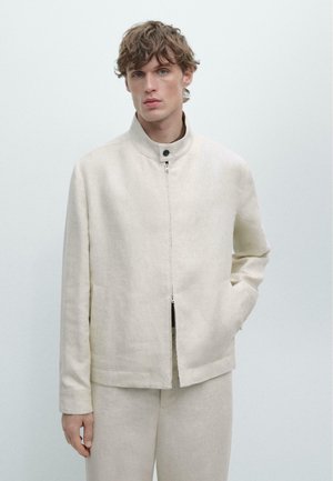 Jeune homme aux cheveux ondulés portant une veste zippée couleur crème et un pantalon assorti, debout avec une main dans la poche sur un fond uni.