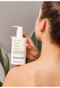 Weiße Lotionflasche mit Pumpspender, grünem und schwarzem Text, 300 ml, in einer Hand gehalten gegen die Haut, mit einer teilweise sichtbaren Pflanze im Hintergrund.
