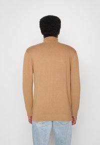 Beige stickad tröja med hög krage, långa ärmar, ribbade muddar och nederkant. Bärs tillsammans med ljusblå jeans, sedd bakifrån.