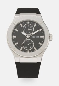 Reloj de esfera negra redonda con acentos plateados, tres subesferas y una correa de silicona negra. Presenta marcadores de hora plateados y un display de fecha.