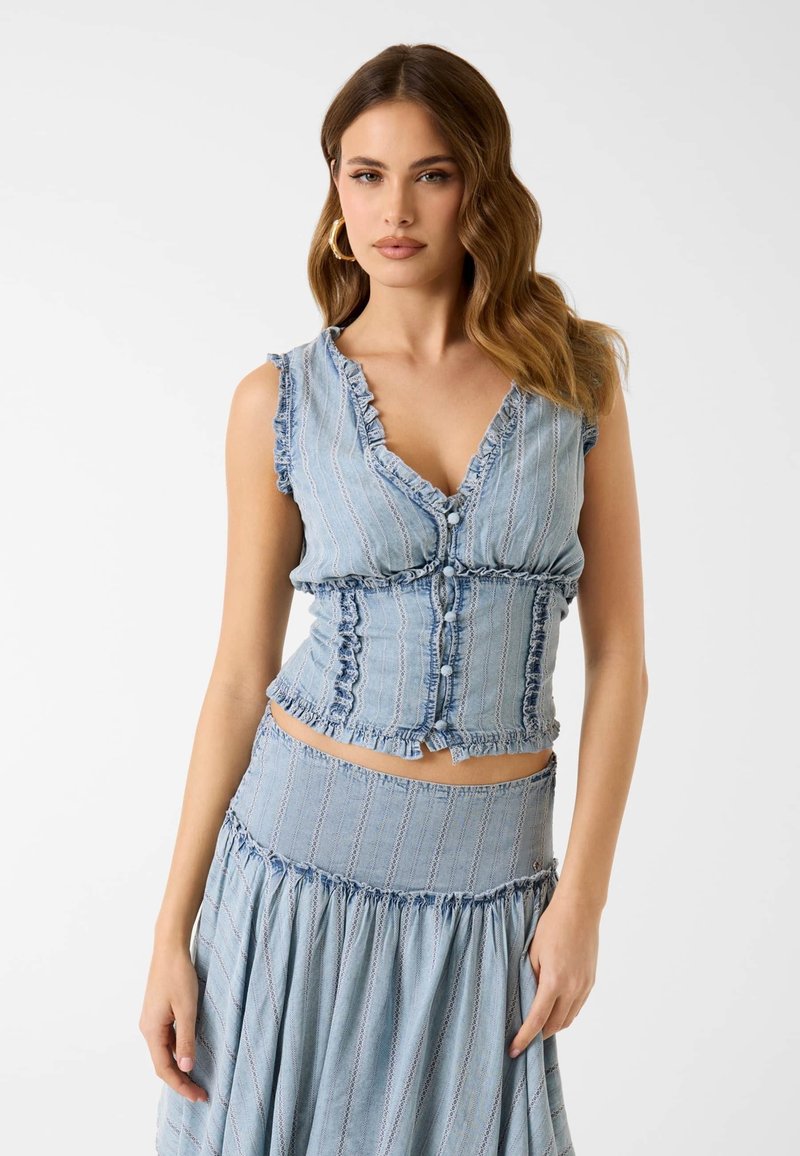 Femme aux cheveux bruns ondulés portant des boucles d’oreilles créoles dorées, un top bleu sans manches à volants et boutons, et une jupe bleue taille haute sur un fond uni.