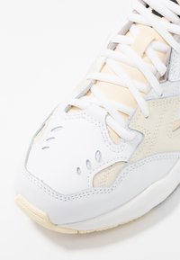 Baskets blanches avec des accents beiges, fabriquées en cuir lisse. Dotées de trous de ventilation perforés, d'une semelle texturée et de lacets plats.