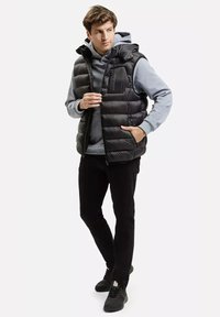 Młody mężczyzna noszący czarną kamizelkę puchową na szarym hoodie, czarne spodnie i czarne sneakersy, stoi z jedną ręką w kieszeni, spoglądając na bok.