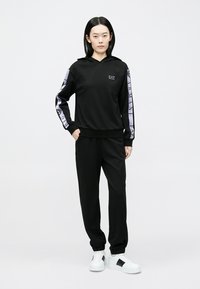 Schwarzer Hoodie mit Logo, langärmlig mit kontrastierendem Streifendesign. Kombiniert mit schwarzen Hosen und weißen Sneakers, glatte Textur.