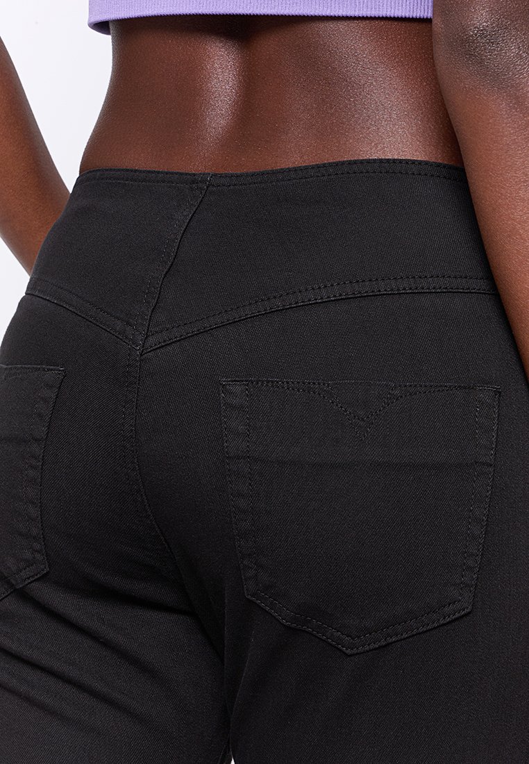 Zbliżenie na osobę w wysokich czarnych dżinsach i krótkim fioletowym topie, ukazujące ich dolną część pleców i biodra z tyłu.