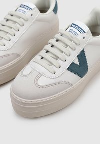 Vita lädersneakers med en texturerad grå mocka tå och grön accent på hälen. Har rund snörning och en tjock gummisul.