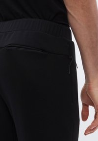 Zbliżenie osoby noszącej czarne spodnie z elastycznym pasem i zamykanym na zamek kieszonką, ręka swobodnie oparta przy boku na jasnym tle.