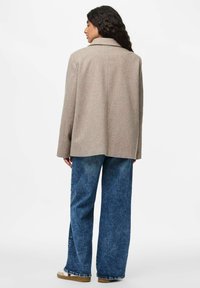 Beež villasegune mantel struktuurse õlavarre, laia varrukaga ja lõikega kraega, millega on kombineeritud lõdvad sinised teksad ja vabaaja kingad.